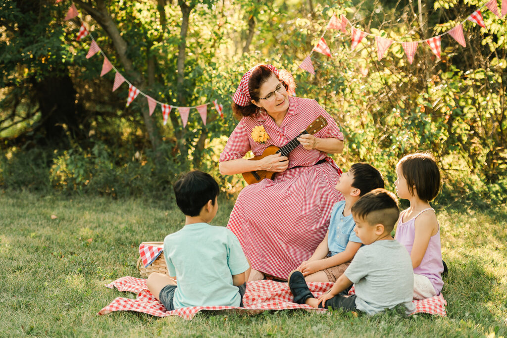 An Edmonton musician in a red gingham dress playing ukelele on a picnic blanket with kids